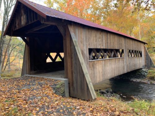 A wooden covered bridge with a red roof, surrounded by autumn foliage and a small stream.