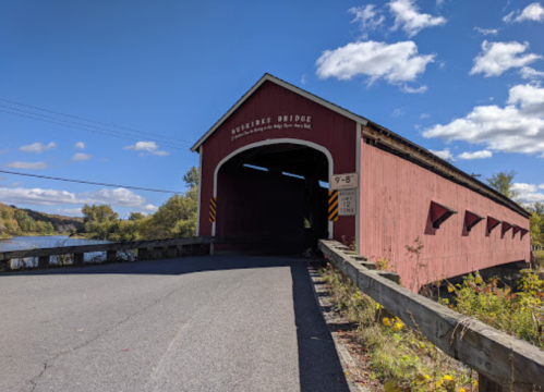 A red covered bridge spans a river under a blue sky with fluffy clouds, surrounded by autumn foliage.