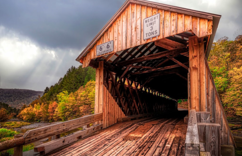 A wooden covered bridge with a weight limit sign, surrounded by autumn foliage and a cloudy sky.