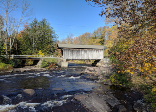 A wooden covered bridge spans a river, surrounded by autumn foliage and clear blue skies.