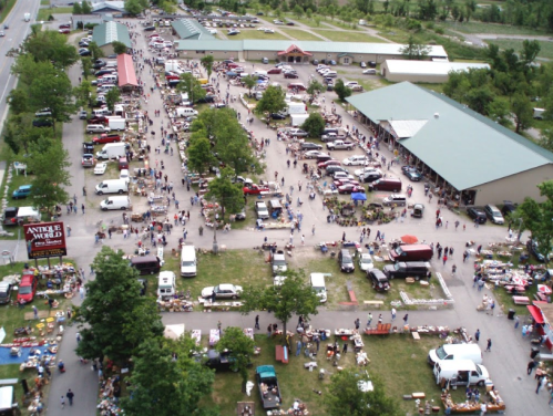 Aerial view of a bustling outdoor market with vendors, shoppers, and parked cars amidst green trees.