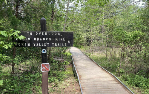 Signpost directing to Overlook, Cabin Branch Mine, and North Valley Trails, with a wooden path surrounded by greenery.