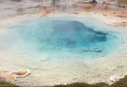 A vibrant blue hot spring surrounded by mineral deposits and steam in a natural landscape.