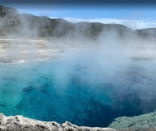 A vibrant blue hot spring surrounded by steam and rocky terrain in a natural landscape.