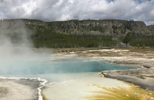 A geothermal pool with vibrant blue water and yellow mineral deposits, surrounded by a misty landscape and green hills.
