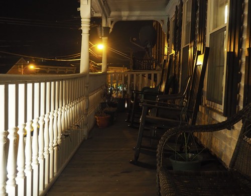 A dimly lit porch at night with rocking chairs and potted plants, illuminated by streetlights in the background.