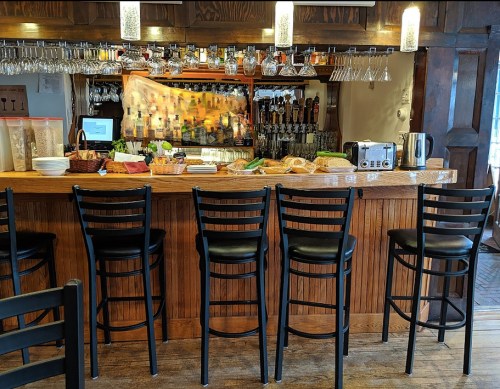 A wooden bar with high stools, bottles on shelves, and a variety of food items displayed on the counter.