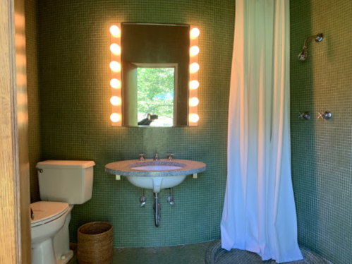A modern bathroom featuring a green tiled wall, a sink, a toilet, a shower with a curtain, and a mirror with lights.