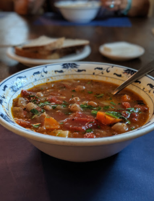 A bowl of hearty vegetable soup with chunks of vegetables and herbs, accompanied by slices of bread in the background.