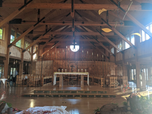 Interior of a wooden church with a high ceiling, altar, and large windows, featuring natural light and a serene atmosphere.