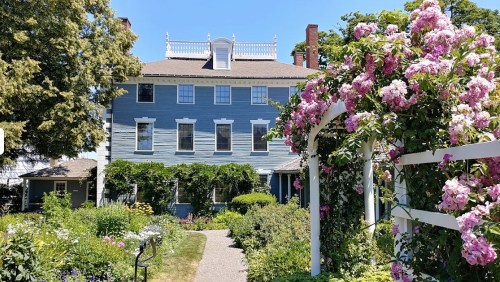 A blue historic house surrounded by lush gardens and blooming pink flowers on a sunny day.