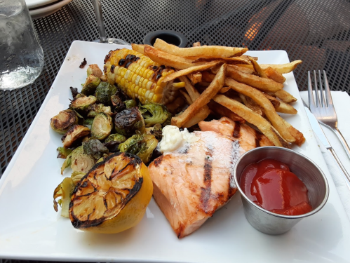 A plate with grilled salmon, corn on the cob, Brussels sprouts, French fries, and a lemon wedge with ketchup on the side.