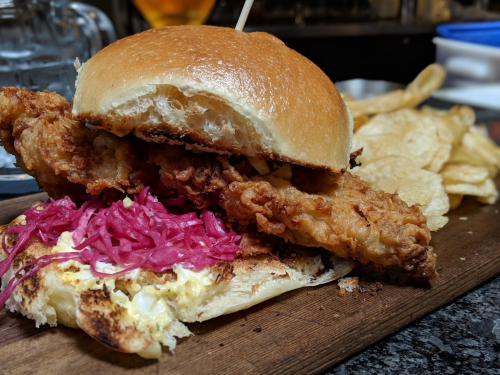 A crispy fried chicken sandwich with pickled red onions on a toasted bun, served with chips and fries on the side.