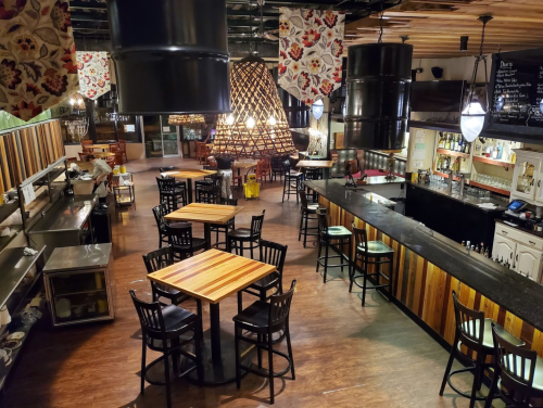 A spacious restaurant interior with wooden tables, black chairs, and decorative hanging lights.
