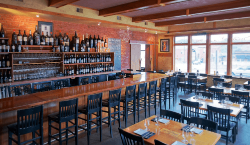 A cozy restaurant interior featuring a wooden bar, wine bottles, and tables with black chairs and natural light.