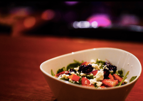 A close-up of a salad in a white bowl, featuring greens, strawberries, blueberries, and feta cheese on a wooden table.