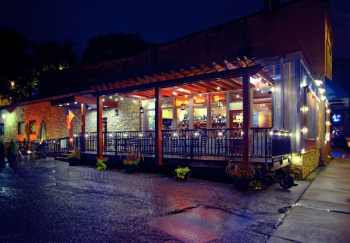 A cozy restaurant exterior at night, featuring a lit patio with seating and a bar, surrounded by plants.