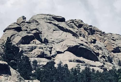 A rugged rock formation rises above a forested area under a cloudy sky.