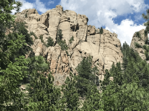 Rocky cliff face surrounded by lush green trees under a partly cloudy blue sky.