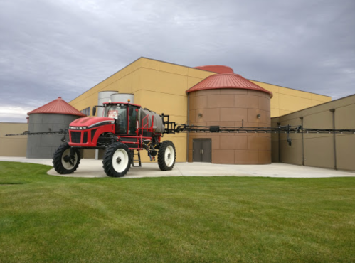A red and black agricultural sprayer parked near large storage silos and a building with a yellow exterior.