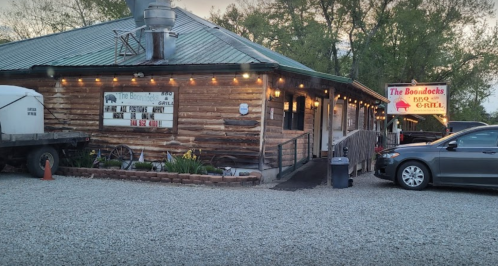 A rustic wooden building with a sign reading "The Boondocks BBQ & Grill," surrounded by gravel and trees.