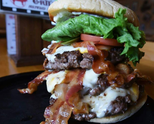 A towering burger with two beef patties, melted cheese, bacon, lettuce, and tomato, served on a bun.