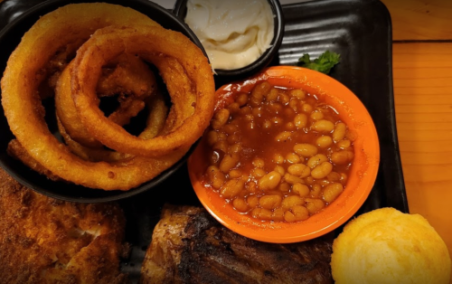 A plate featuring onion rings, baked beans, cornbread, and a side of creamy dip.