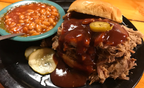 A plate featuring a pulled pork sandwich topped with barbecue sauce and pickles, alongside a bowl of baked beans.