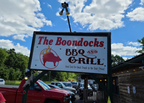 Sign for "The Boondocks BBQ and Grill" with a cow graphic, under a blue sky with clouds. Red trucks parked nearby.