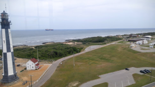 A coastal view featuring a lighthouse, grassy area, and ocean with a ship in the distance under a cloudy sky.