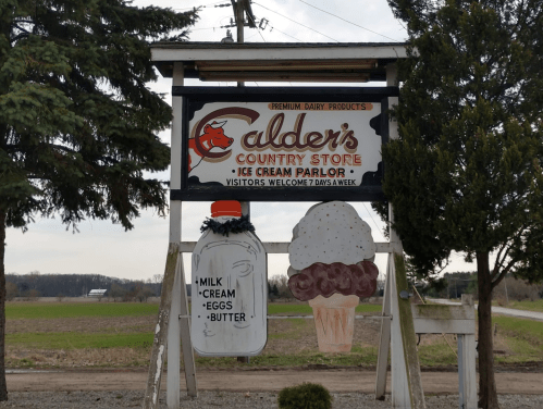 Sign for Calder's Country Store and Ice Cream Parlor, featuring a milk bottle and ice cream cone, surrounded by trees.