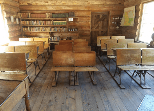 A rustic classroom with wooden desks, shelves of books, and a door, set in a log cabin.