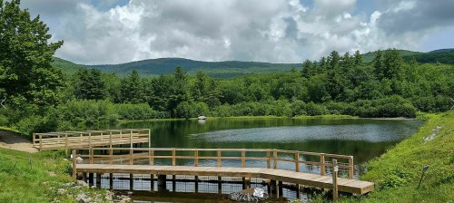 A serene landscape featuring a calm pond surrounded by lush greenery and distant mountains under a partly cloudy sky.