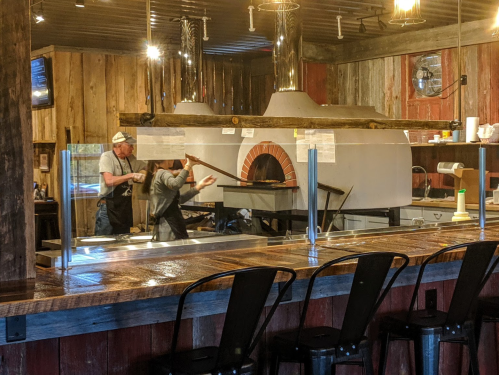 A rustic kitchen with a wood-fired oven, chefs preparing food, and a bar area with metal stools.