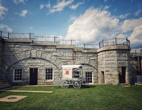 Historic stone building with a white wagon in front, set against a blue sky with clouds. Green grass in the foreground.