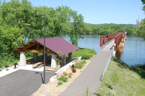 A scenic view of a riverside pavilion and bridge surrounded by greenery on a sunny day.