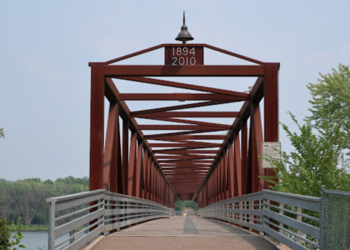 A red metal bridge spans a river, with the years 1894 and 2010 displayed above the entrance. Lush greenery surrounds it.