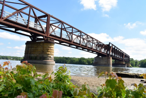 A rusted metal bridge spans a river, supported by stone pillars, with greenery in the foreground and a blue sky above.