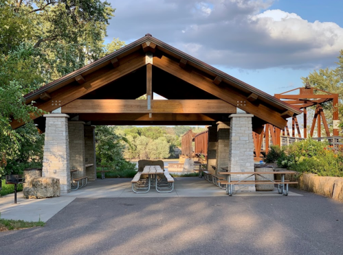 A wooden pavilion with stone columns and picnic tables, surrounded by greenery and a pathway.