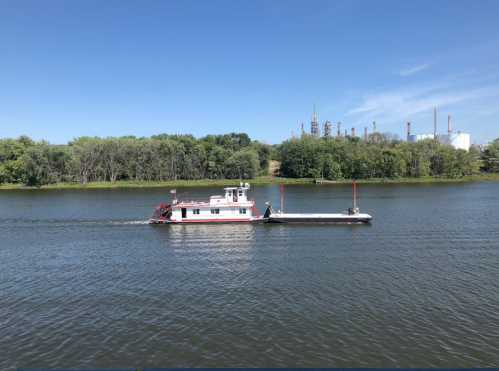 A pink and white boat navigates a calm river, surrounded by green trees and industrial buildings in the background.