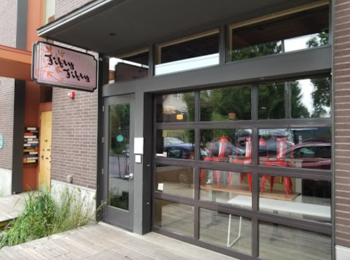 Exterior view of a restaurant with large glass doors and a sign reading "Jitty Jitty," surrounded by greenery.