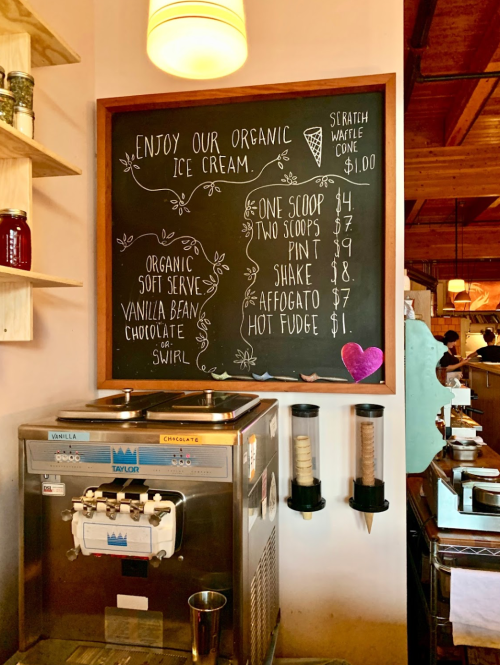 Chalkboard menu displaying organic ice cream options and prices next to a soft serve machine.