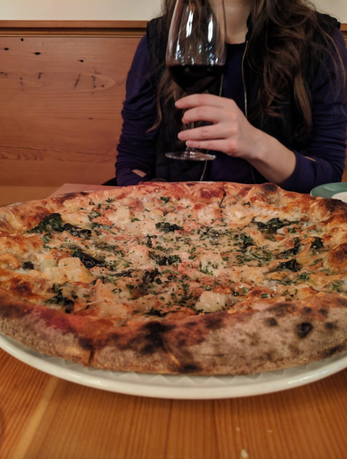 A close-up of a pizza topped with greens and cheese, with a person holding a glass of red wine in the background.