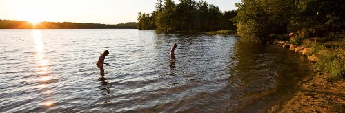 Two children wade in a calm lake at sunset, surrounded by trees and a serene landscape.