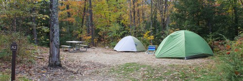 Two tents set up in a wooded campsite with a picnic table and chairs, surrounded by autumn foliage.