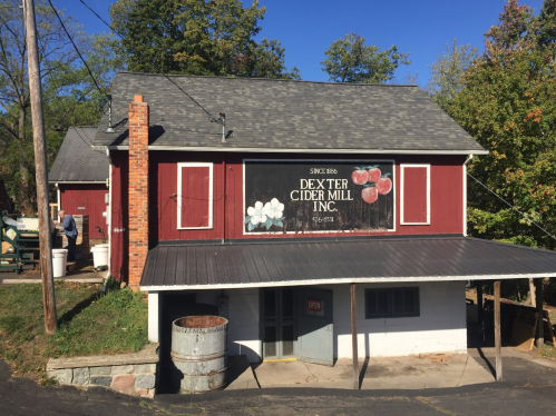 Red barn-style building with a sign reading "Dexter Cider Mill Inc." and an open door, surrounded by trees.