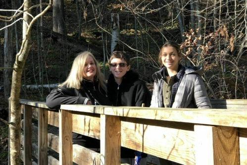 Three people smiling on a wooden bridge in a forested area, surrounded by trees and nature.