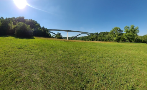 A wide green field with a modern bridge spanning across it, surrounded by trees under a clear blue sky.