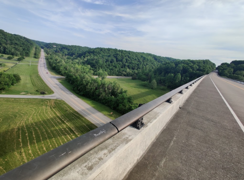 A scenic view from a bridge overlooking green hills and winding roads under a clear sky.