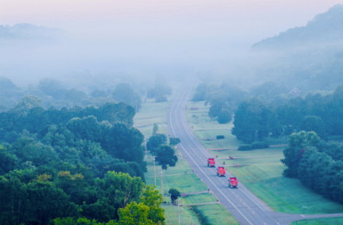 A misty morning view of a road with three red trucks traveling through a green landscape.